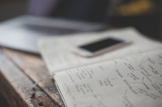 An open notebook with handwritten notes is placed on a wooden table next to a smartphone. The notes appear to focus on personal development and creative values. A partially visible laptop is in the background, creating a workspace setting.