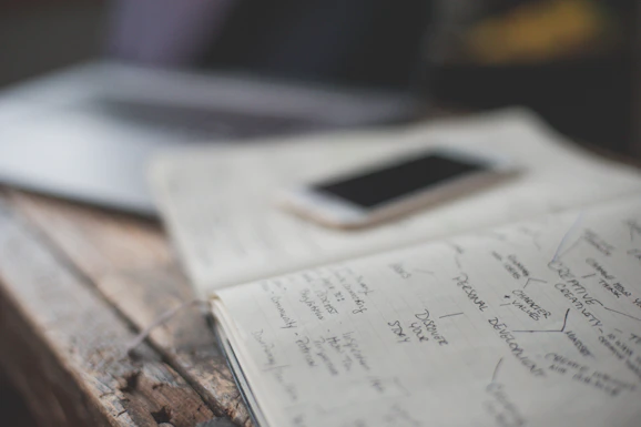 An open notebook with handwritten notes is placed on a wooden table next to a smartphone. The notes appear to focus on personal development and creative values. A partially visible laptop is in the background, creating a workspace setting.
