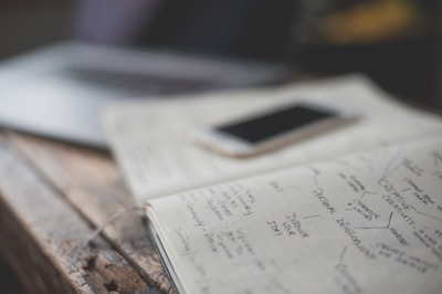 An open notebook with handwritten notes is placed on a wooden table next to a smartphone. The notes appear to focus on personal development and creative values. A partially visible laptop is in the background, creating a workspace setting.