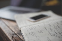 An open notebook with handwritten notes is placed on a wooden table next to a smartphone. The notes appear to focus on personal development and creative values. A partially visible laptop is in the background, creating a workspace setting.