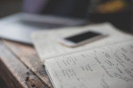An open notebook with handwritten notes is placed on a wooden table next to a smartphone. The notes appear to focus on personal development and creative values. A partially visible laptop is in the background, creating a workspace setting.
