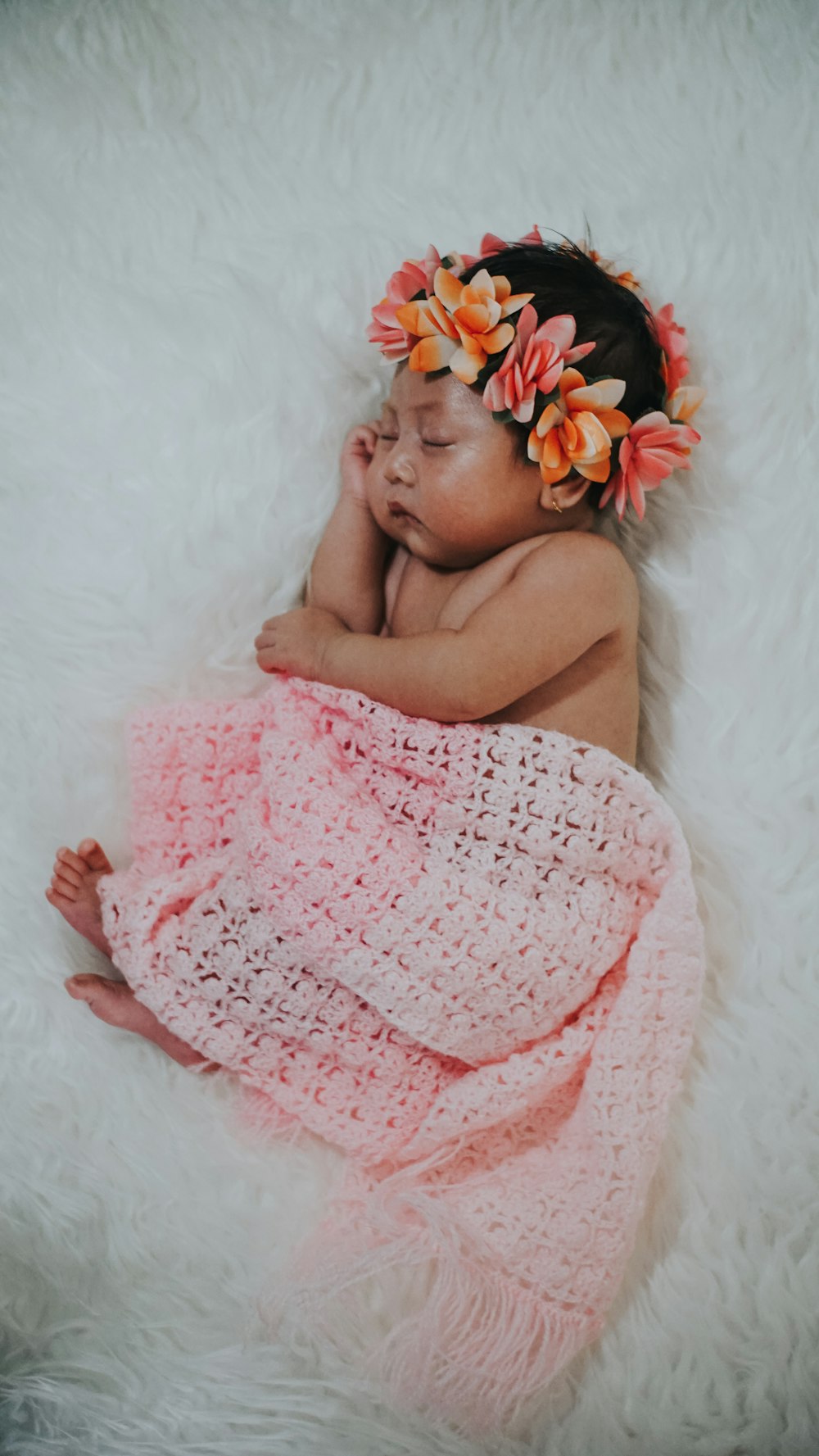 bébé porte une couronne florale et est recouvert d’un châle rose