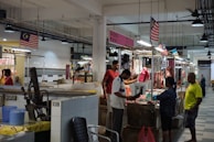An indoor market scene where several people are interacting at stalls displaying various cuts of meat. The stalls are well-lit, with overhead lights hanging from the ceiling, and Malaysian flags are visible throughout the area. The setting is clean, with tiled floors and walls. A man in a white shirt appears to be cutting meat on a wooden block, while others look on or engage in conversation.