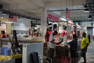 An indoor market scene where several people are interacting at stalls displaying various cuts of meat. The stalls are well-lit, with overhead lights hanging from the ceiling, and Malaysian flags are visible throughout the area. The setting is clean, with tiled floors and walls. A man in a white shirt appears to be cutting meat on a wooden block, while others look on or engage in conversation.