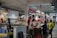 An indoor market scene where several people are interacting at stalls displaying various cuts of meat. The stalls are well-lit, with overhead lights hanging from the ceiling, and Malaysian flags are visible throughout the area. The setting is clean, with tiled floors and walls. A man in a white shirt appears to be cutting meat on a wooden block, while others look on or engage in conversation.