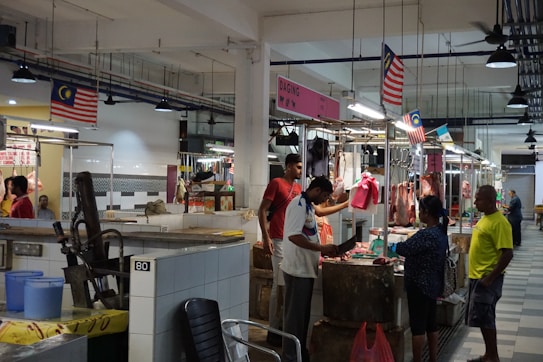 An indoor market scene where several people are interacting at stalls displaying various cuts of meat. The stalls are well-lit, with overhead lights hanging from the ceiling, and Malaysian flags are visible throughout the area. The setting is clean, with tiled floors and walls. A man in a white shirt appears to be cutting meat on a wooden block, while others look on or engage in conversation.