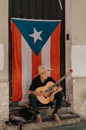 A person sits on a step with a guitar, wearing a straw hat with a small flag emblem on it. Behind them is a large Puerto Rican flag hanging on a door. The individual appears to be resting, adding a sense of calm to the setting.