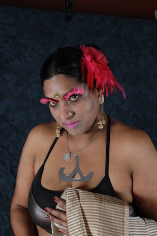 A person with vibrant makeup, including pink feather eyelashes and pink lipstick, stands against a dark background. They wear a metal pendant necklace and hold a striped fabric over their shoulder. A red feathered hair accessory and dangling earrings complete the look.