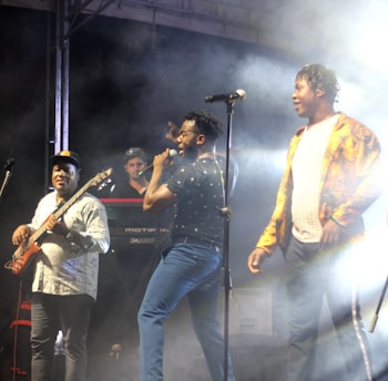 A lively performance on stage features three musicians. One is playing a bass guitar, another is singing into a microphone, and the third stands next to them. The background includes a keyboard and stage lighting creating a smoky atmosphere.