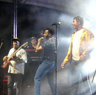 A lively performance on stage features three musicians. One is playing a bass guitar, another is singing into a microphone, and the third stands next to them. The background includes a keyboard and stage lighting creating a smoky atmosphere.