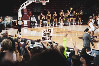 A lively basketball game with fans in the foreground holding a sign that reads 'Make Some Noise'. The scene captures a court with players in action, surrounded by enthusiastic cheerleaders in the background. A referee is also visible, adding to the excitement of the game.