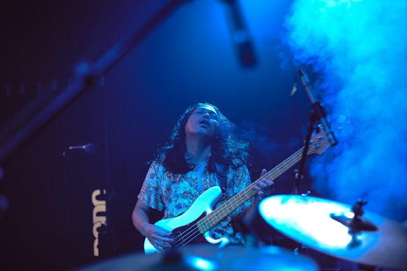A musician passionately playing an electric bass guitar on stage, surrounded by blue stage lights and smoke. The person has long hair and is wearing a patterned shirt. The scene includes a drum set and microphones, indicating a live performance.