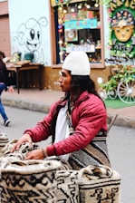 A person wearing a white knit cap and a red quilted jacket is standing by a display of woven bags with intricate patterns. The background includes a colorful store window and graffiti art on the wall. The street setting is lively and vibrant.