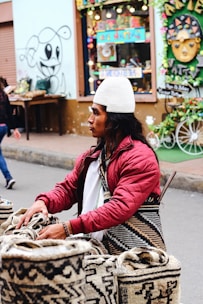 A person wearing a white knit cap and a red quilted jacket is standing by a display of woven bags with intricate patterns. The background includes a colorful store window and graffiti art on the wall. The street setting is lively and vibrant.