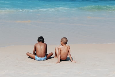 two topless children sitting near seashore during daytime back teams background