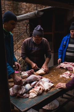 Several individuals are gathered around a wooden table outdoors, engaged in cutting large pieces of raw meat. The setting appears to be rustic, with a brick wall and metal railings in the background. The people are dressed warmly, suggesting a cold environment.