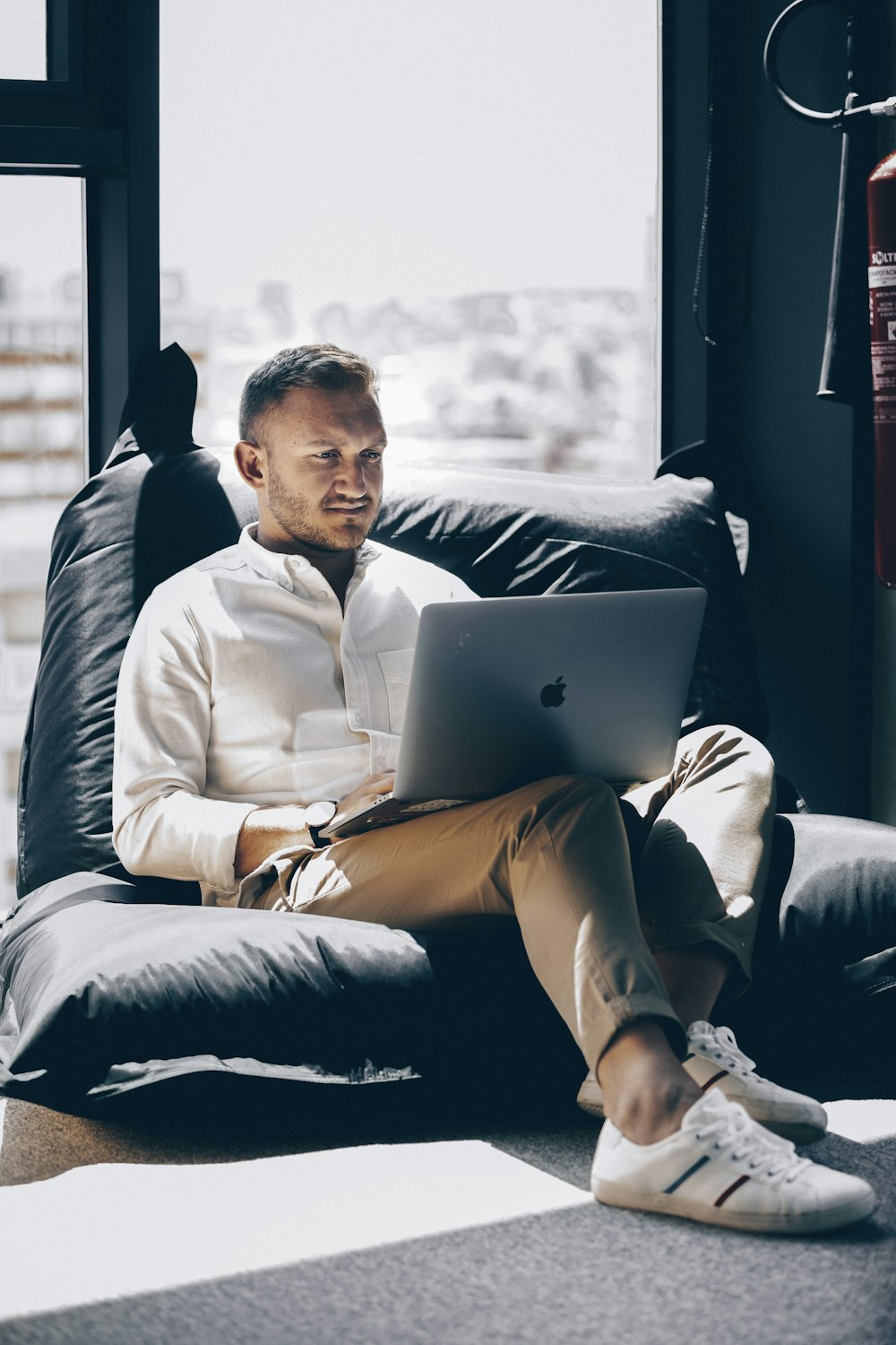 man sitting on bed chair while using silver MacBook near window during daytime