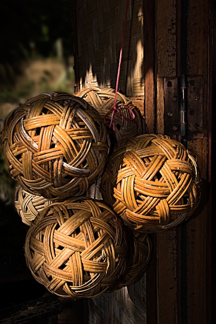 A collection of woven rattan balls hangs from a red cord against a background of woven wooden panels. The sunlight creates a play of shadows and highlights on the textured surface of the rattan and the wooden backdrop, enhancing the warm tones and natural patterns.