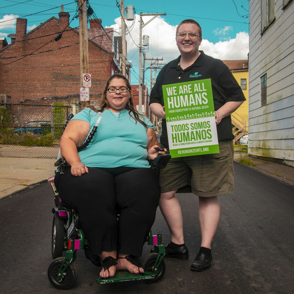 woman sitting on wheelchair beside man holding we are all humans sign