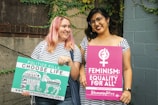 Two people stand side by side in front of a brick and concrete wall with ivy. Both hold protest signs. One holds a green and white sign that says 'Politics Kills. Choose Life.' and has various words related to political topics. The other holds a pink sign stating 'Feminism: Equality For All.' Both individuals are smiling and have casual attire, with striped tops and glasses.
