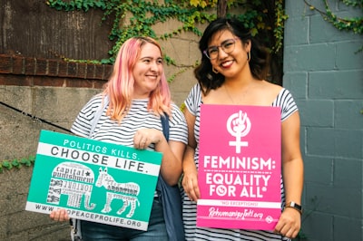 two smiling women standing while holding banners near concrete wall republican zoom background