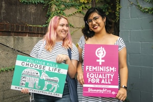 Two people stand side by side in front of a brick and concrete wall with ivy. Both hold protest signs. One holds a green and white sign that says 'Politics Kills. Choose Life.' and has various words related to political topics. The other holds a pink sign stating 'Feminism: Equality For All.' Both individuals are smiling and have casual attire, with striped tops and glasses.
