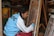 A person is focused on painting a colorful intricate design on a canvas, wearing a blue and white jacket with 'MCFC' on the back. The scene is set in what appears to be an artist's workshop, with various art supplies and other framed artworks visible.