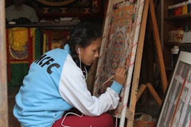 A person is focused on painting a colorful intricate design on a canvas, wearing a blue and white jacket with 'MCFC' on the back. The scene is set in what appears to be an artist's workshop, with various art supplies and other framed artworks visible.