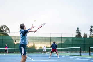 A group of four people engaging in a doubles tennis match on an outdoor court. The focus is on the player in the foreground serving, with a tennis racket in hand, preparing to hit the ball. The players are on a blue tennis court surrounded by a green fence, with trees visible in the background. The lighting suggests it might be early morning or late afternoon.