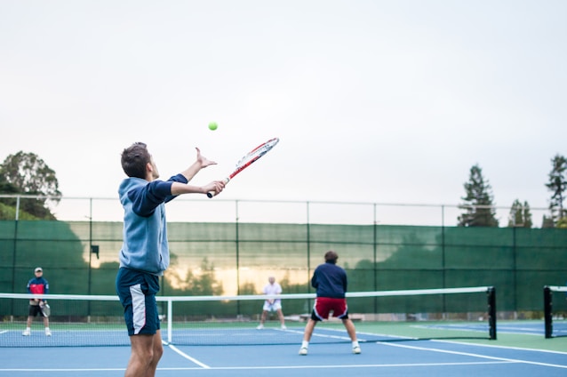 A group of four people engaging in a doubles tennis match on an outdoor court. The focus is on the player in the foreground serving, with a tennis racket in hand, preparing to hit the ball. The players are on a blue tennis court surrounded by a green fence, with trees visible in the background. The lighting suggests it might be early morning or late afternoon.