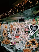 A festive market stall filled with an array of Christmas-themed decorations and ornaments. The display includes baskets of dried oranges, plush toys, and various hanging trinkets. The whole setup is illuminated by warm, twinkling fairy lights. A chalkboard sign reads 'We Accept Credit Cards'. Evergreen garlands add to the holiday atmosphere.