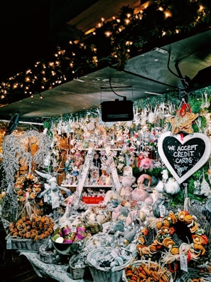 A festive market stall filled with an array of Christmas-themed decorations and ornaments. The display includes baskets of dried oranges, plush toys, and various hanging trinkets. The whole setup is illuminated by warm, twinkling fairy lights. A chalkboard sign reads 'We Accept Credit Cards'. Evergreen garlands add to the holiday atmosphere.