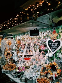A festive market stall filled with an array of Christmas-themed decorations and ornaments. The display includes baskets of dried oranges, plush toys, and various hanging trinkets. The whole setup is illuminated by warm, twinkling fairy lights. A chalkboard sign reads 'We Accept Credit Cards'. Evergreen garlands add to the holiday atmosphere.