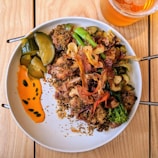 A plate of food featuring roasted chicken topped with crispy vegetables alongside pickles and a vibrant orange sauce. The dish is garnished with herbs, accompanied by a glass of amber-colored drink.