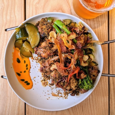 A plate of food featuring roasted chicken topped with crispy vegetables alongside pickles and a vibrant orange sauce. The dish is garnished with herbs, accompanied by a glass of amber-colored drink.