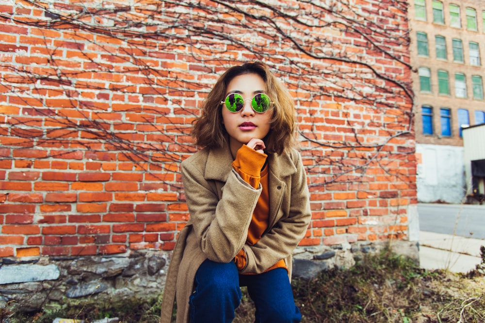 woman wearing brown zip-up jacket and green framed sunglasses near orange bricked wall