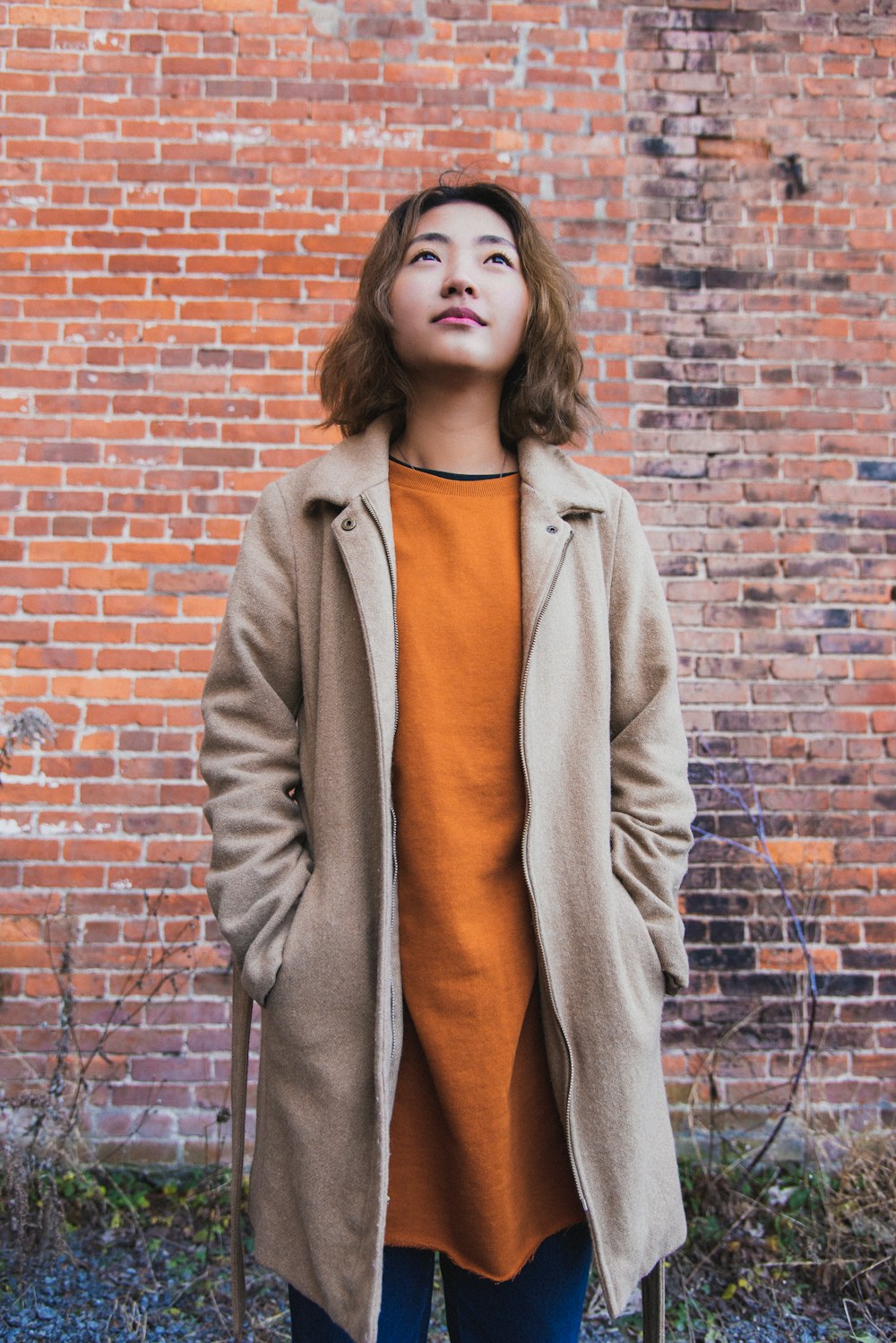Mujer con camisa naranja y abrigo beige de pie cerca de la pared de ladrillo