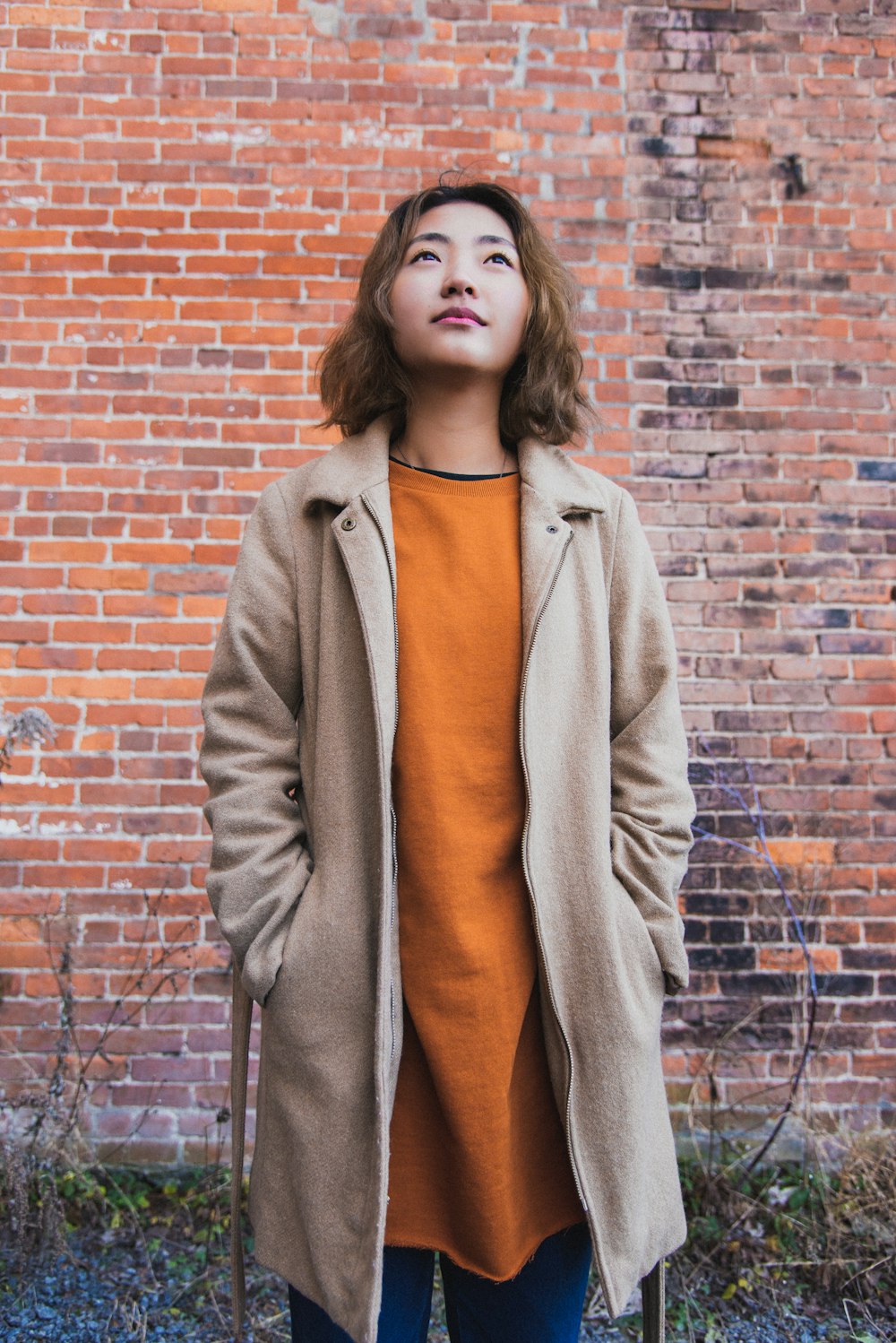 woman wearing orange shirt and beige coat standing near brick wall