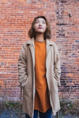 woman wearing orange shirt and beige coat standing near brick wall