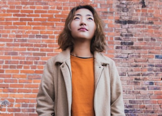 woman wearing orange shirt and beige coat standing near brick wall