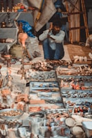 A man sits on the ground surrounded by an array of items displayed for sale, including metal cookware, wooden objects, and various handcrafted goods. The setting appears to be a market stall with vibrant textiles and wooden structures in the background.
