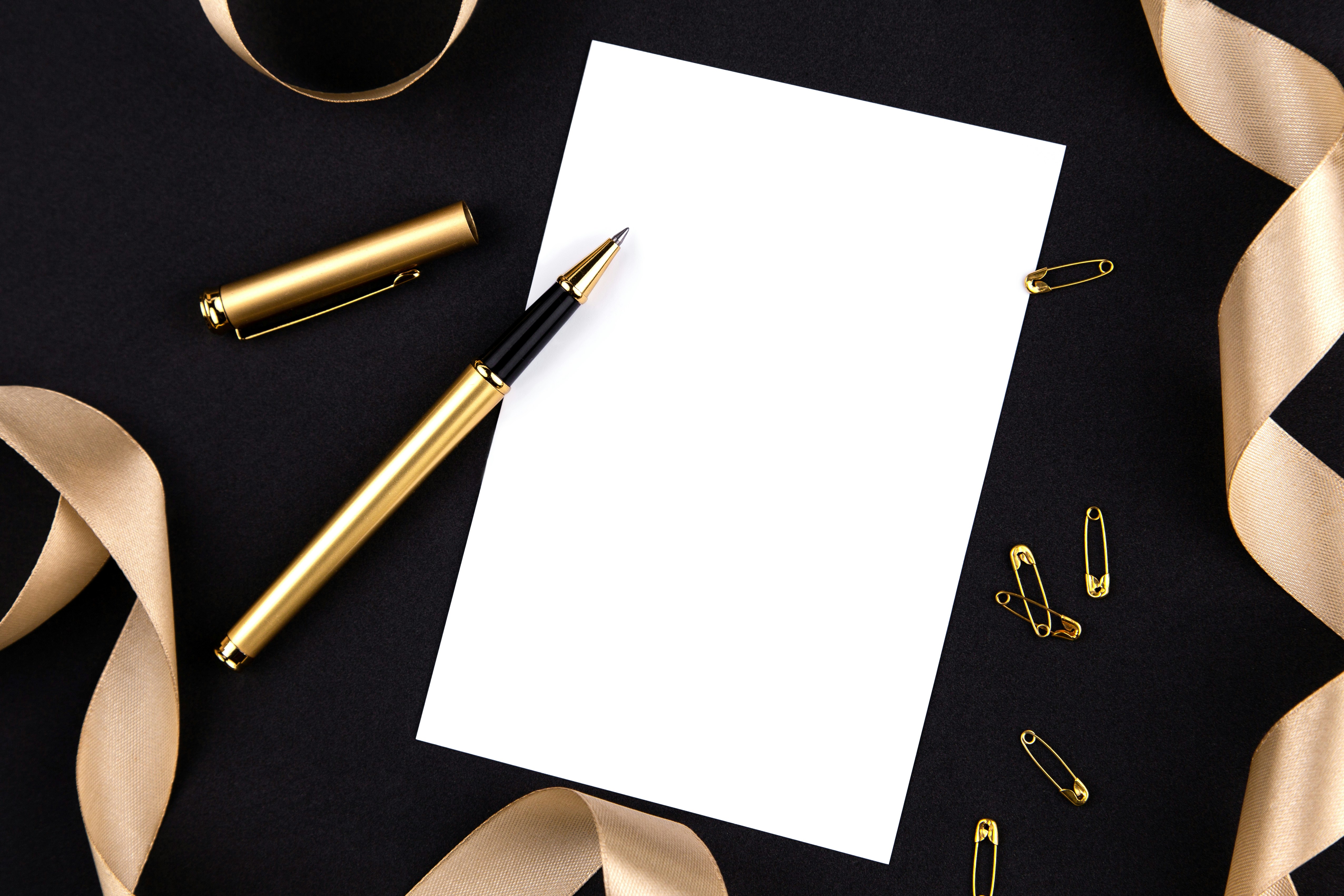 Gold pen, ribbon, paper clips and stationery on a black background with a white sheet of paper.