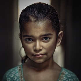 A young girl with a serious expression stands in soft lighting, highlighting her facial features. She has dark hair and is wearing a green garment with intricate designs. The background is softly blurred, focusing attention on her face.