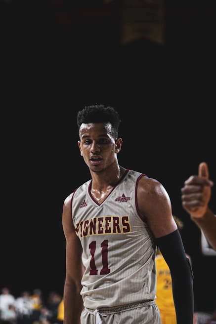 A basketball player wearing a white and maroon 'Pioneers' jersey with the number 11 is standing on a court. The background is dark with blurred figures and possibly another player in a yellow jersey. The player appears focused and slightly sweaty.