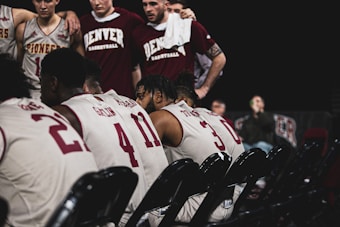 A group of basketball players wearing jerseys with numbers are seated in rows on folding chairs. In the background, other team members stand close together, some with arms around each other, in a supportive manner. The jerseys have the words 'Pioneers' and 'Denver Basketball' visible. The setting seems to be an indoor sports arena.