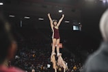 A cheerleader is lifted high into the air by her teammates, forming a human pyramid. She has her arms raised triumphantly, smiling under the spotlights of an indoor sports arena. The audience sits in the background, observing the performance.