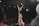 A cheerleader is lifted high into the air by her teammates, forming a human pyramid. She has her arms raised triumphantly, smiling under the spotlights of an indoor sports arena. The audience sits in the background, observing the performance.