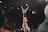 A cheerleader is lifted high into the air by her teammates, forming a human pyramid. She has her arms raised triumphantly, smiling under the spotlights of an indoor sports arena. The audience sits in the background, observing the performance.