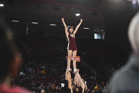 A cheerleader is lifted high into the air by her teammates, forming a human pyramid. She has her arms raised triumphantly, smiling under the spotlights of an indoor sports arena. The audience sits in the background, observing the performance.