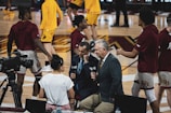 A group of individuals, likely basketball players, is on a gymnasium court. Some are wearing yellow athletic outfits, and others are in burgundy athletic gear. Two men in suits are speaking into microphones while a woman stands nearby wearing a white shirt. A camera setup is positioned to capture the scene, indicating a broadcast or interview setting.